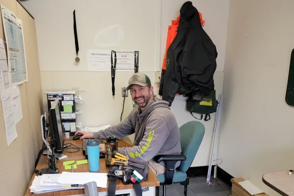 A Peninsula College worker sits at a desk next to a tool belt. He is smiling, wearing a grey Carhartt hoodie.