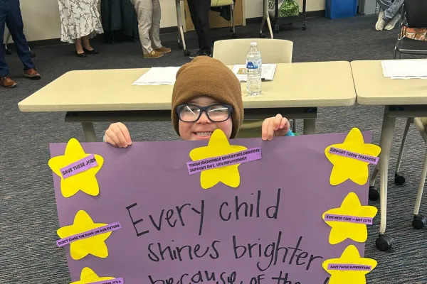 A child holds a sign reading "Every child shines brighter because of them!"