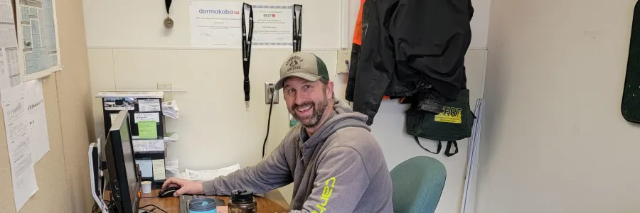A Peninsula College worker sits at a desk next to a tool belt. He is smiling, wearing a grey Carhartt hoodie.