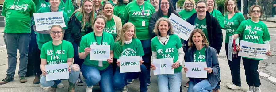 WFSE members in green shirts stand on a street corner and hold signs that write out "SUPPORT STAFF SUPPORT US ALL"