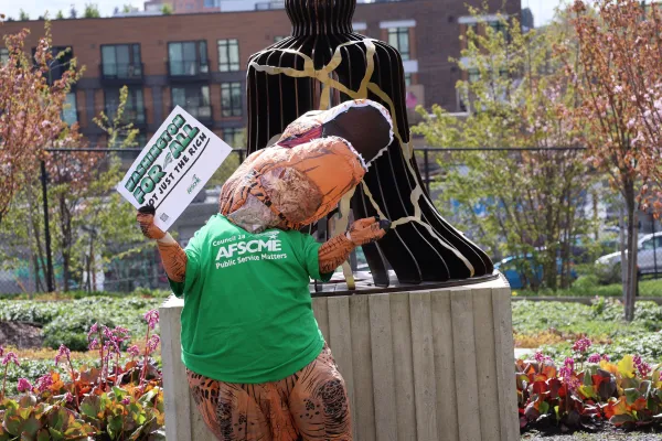 A WFSE member in a t-rex costume in a WFSE shirt holds a sign saying "Washington for all, not just the rich"