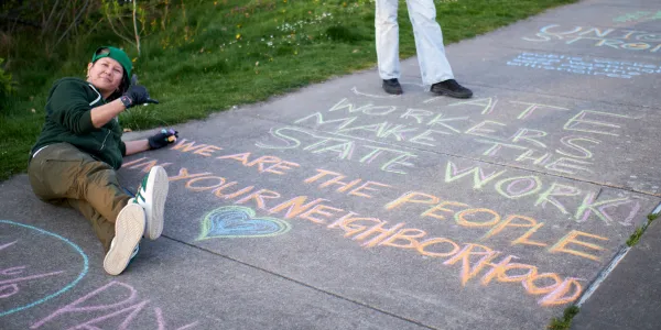 A WFSE member poses next to chalk art stating "State workers make the state work. We are the people in your neighborhood"